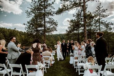 A newlywed couple walks down the aisle outdoors with guests clapping around them.