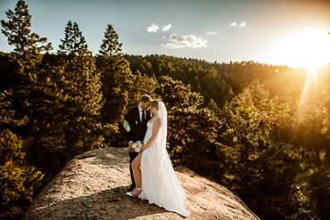 Newlyweds embrace on a sunlit rock surrounded by forest.