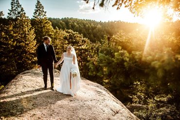 A couple in wedding attire walking hand-in-hand on a sunlit rocky hill surrounded by pine trees.