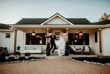 A joyful bride and groom jump in front of a charming house after their wedding.