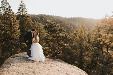 A couple in wedding attire kissing on a sunlit rock surrounded by pine trees.