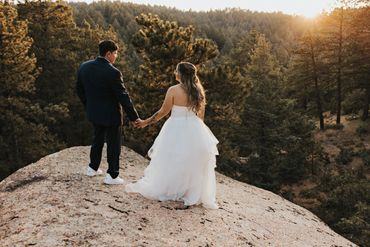 Newlyweds hold hands on a rocky overlook at sunset surrounded by pine trees.