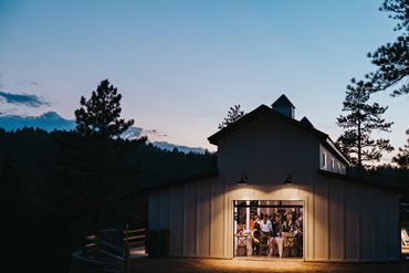 A warmly lit barn hosting a gathering at dusk in a forested area.