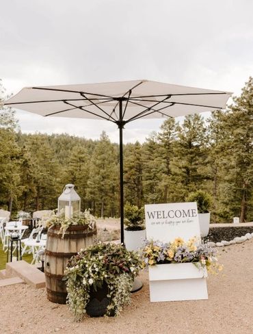 Rustic outdoor welcome setup with umbrella, flowers, and wooden barrel.