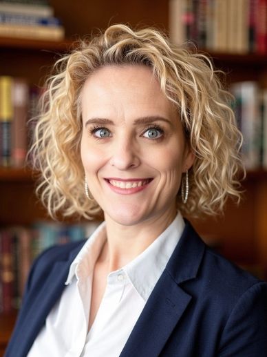 Professional woman with curly blonde hair smiles in front of bookshelves.