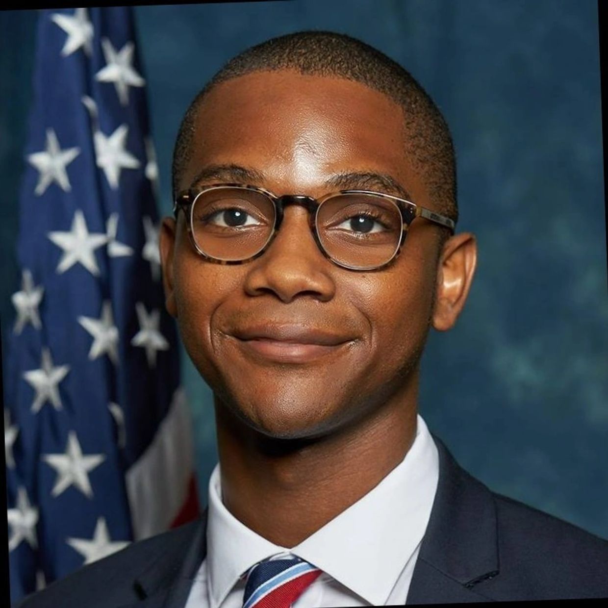Professional portrait of a smiling man with glasses, wearing a suit and tie, with an American flag in the background.