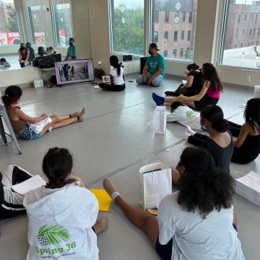 Group of people sitting on the floor watching a screen in a bright room.