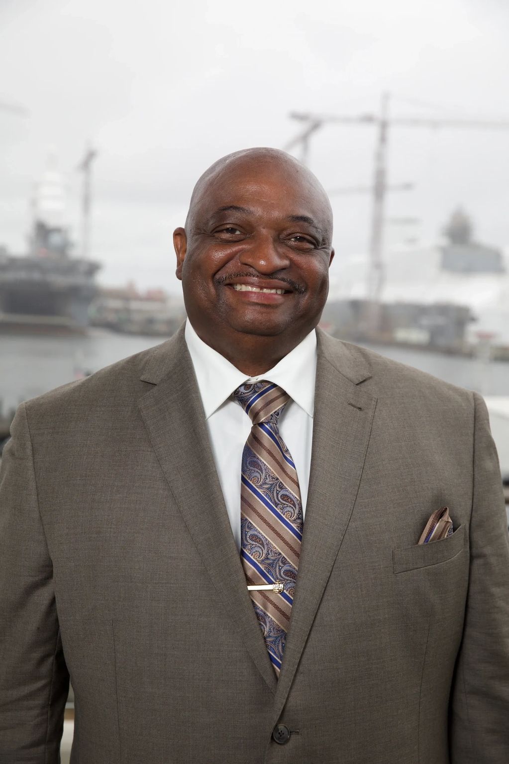 Smiling man in a suit with a patterned tie, standing outdoors near a harbor.