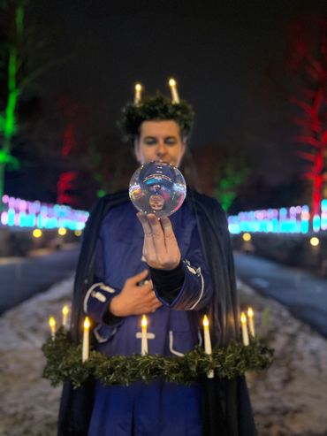A crystal ball juggler wearing a candlelit wreath on his head and another larger one on his waist