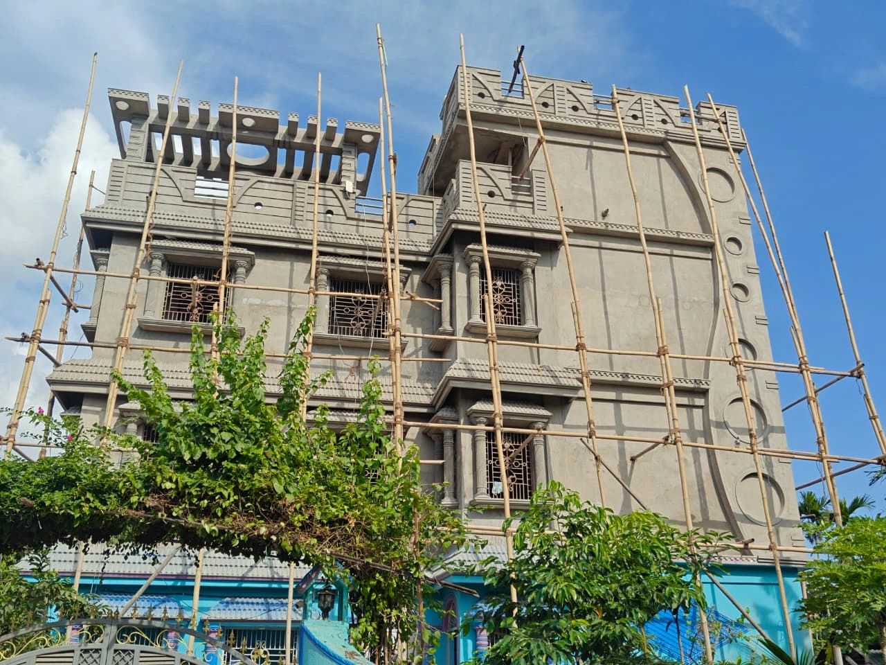 A multi-story building under construction with bamboo scaffolding and greenery in front.