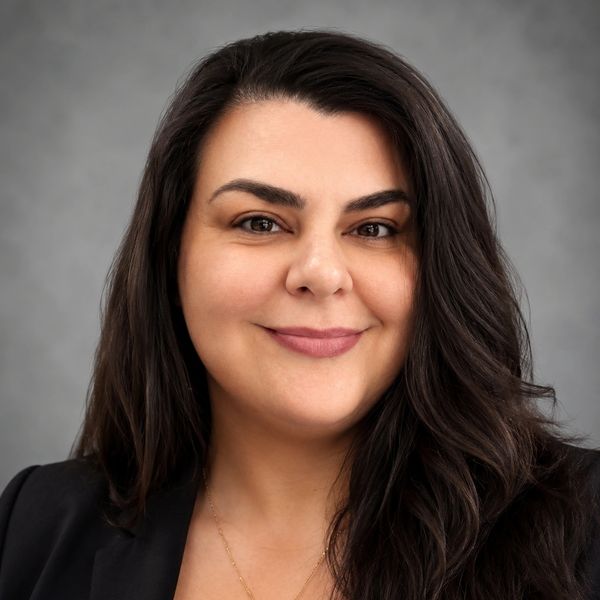 Professional headshot of a smiling woman with long dark hair and a black blazer.