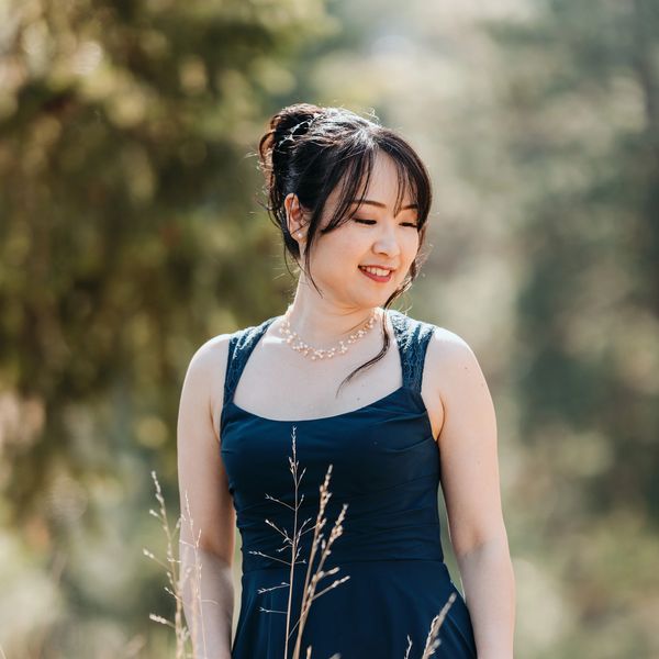Woman in a dark blue dress smiling outdoors with blurred greenery background.
