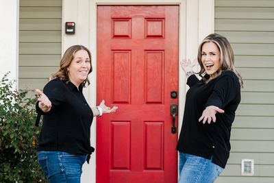 Two women joyfully presenting a red front door of a house.