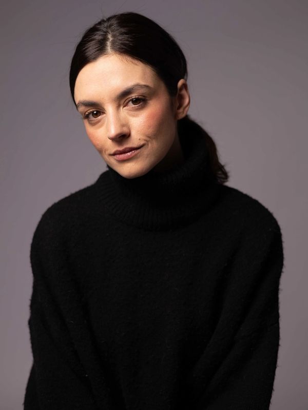 Young woman with dark hair in a black turtleneck sweater against a gray background.