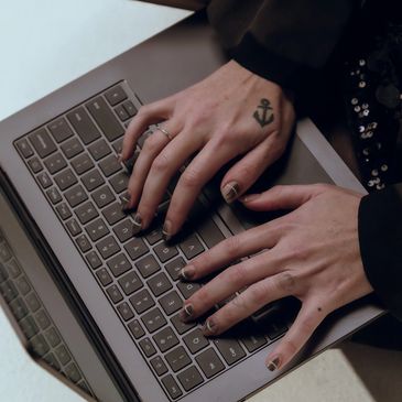 Hands with nail art and anchor tattoo typing on a laptop keyboard.