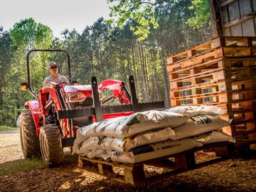 A farmer transports bags of fertilizer on a red tractor into a barn.