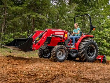 A woman operates a red tractor on a dirt field surrounded by trees.