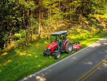 A red tractor mowing grass beside a forest road on a sunny day.
