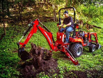 Man operating a red Massey Ferguson mini excavator in a forest clearing.