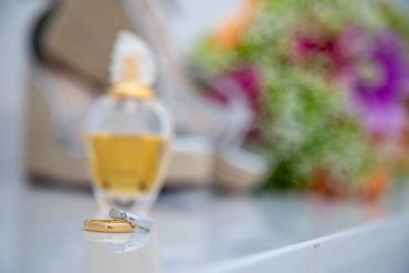 Two wedding rings on a shiny surface with blurred flowers and perfume in the background.