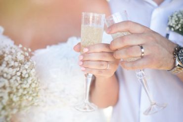 Bride and groom clinking champagne glasses with wedding rings visible.