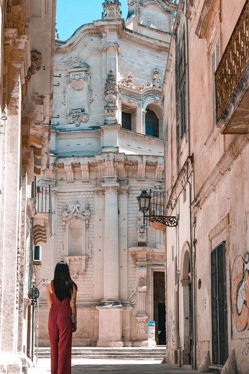 A woman in a red jumpsuit stands in a narrow European street, gazing up at a grand Baroque-style church façade adorned with ornate carvings and columns, bathed in soft sunlight. A woman in a red jumpsuit stands in a narrow European street, gazing up at a grand Baroque-style church façade adorned with ornate carvings and columns, bathed in soft sunlight.