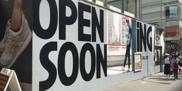 Large black letters on a white wall announce an upcoming store opening.