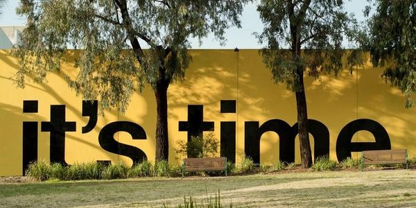 A yellow wall with large black letters saying "it's time" and two trees casting shadows.