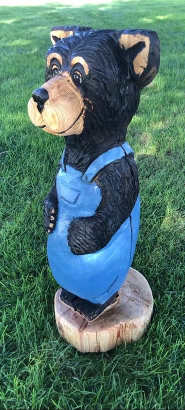 Wooden bear sculpture wearing blue overalls on grass.