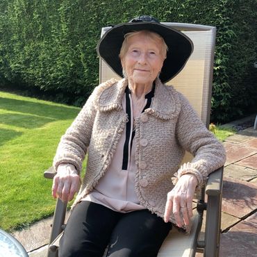 Elderly woman sitting outdoors on a chair, wearing a hat and cozy sweater.