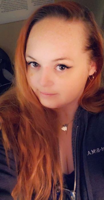 A woman with long red hair wearing a dark jacket and necklace.