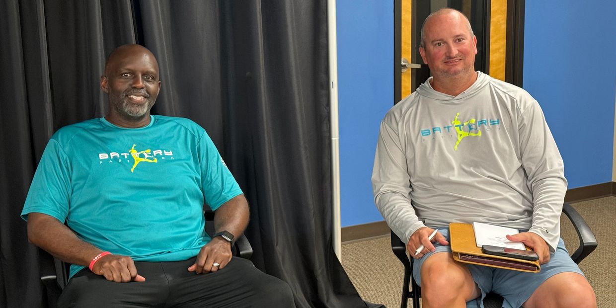 Two men sitting indoors wearing Battery Passion shirts.