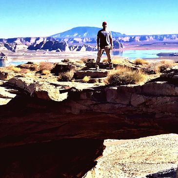 Man standing atop Skylight Arch on tour with Lake Powell and Navajo Mountain behind.