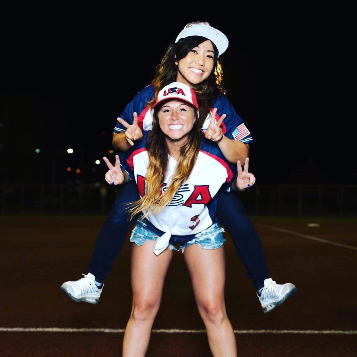 Two women in USA sports jerseys posing happily with peace signs at night.