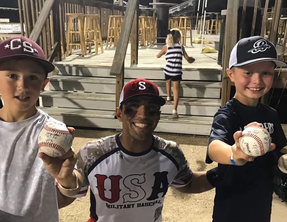 Three smiling baseball players showing signed baseballs, with a girl walking up the stairs in the background.
