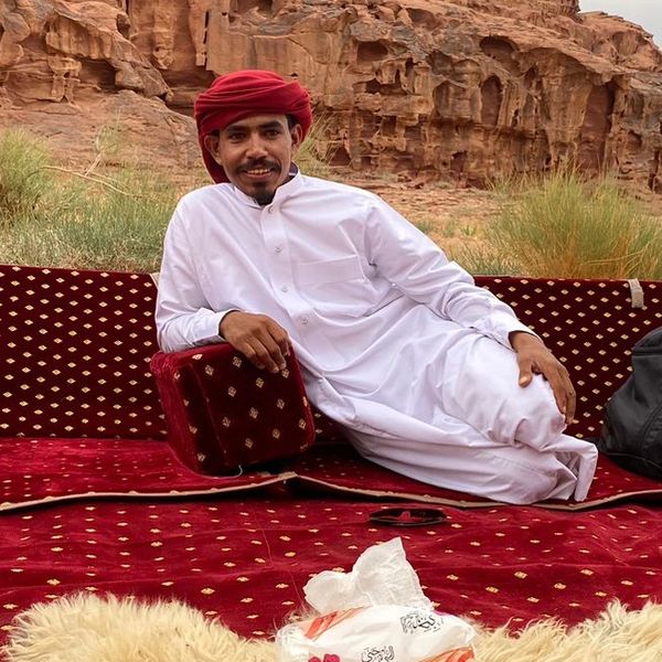 Man in traditional attire lounging on red cushions in a desert landscape.