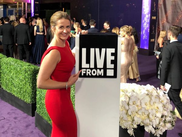 Woman in Red Dress standing on purple carpet pointing to a Live From E! sign