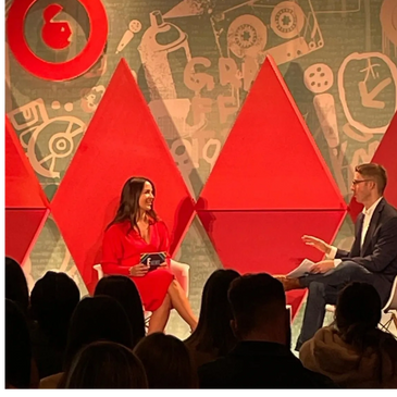 Woman sitting on stage speaking to a man in front of a large crowd