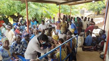 A large group of people gathered outdoors under a shelter, some wearing masks and receiving medical attention.