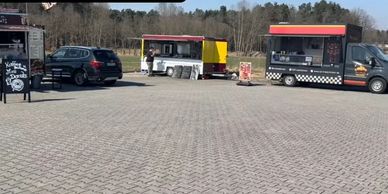 Three food trucks parked with a car in a paved lot near a wooded area.