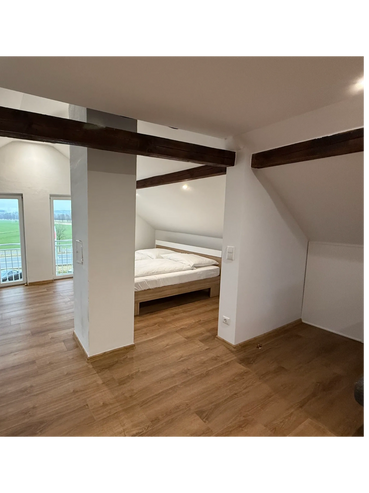 Bright attic bedroom with wooden beams and large windows overlooking a green field.