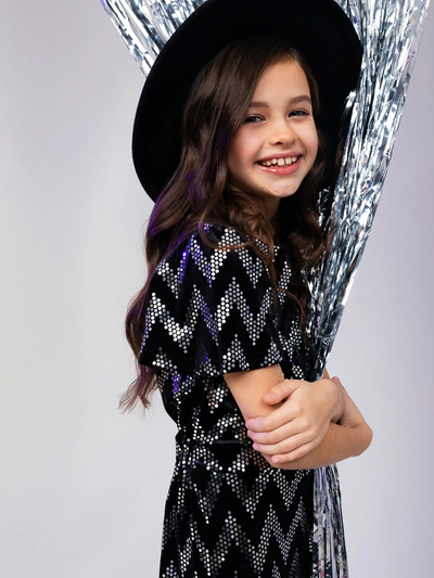 Smiling girl in a black sequined dress and hat holding silver streamers.