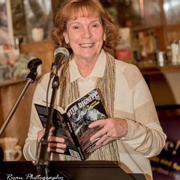 A smiling woman reads from a book at a microphone in a cozy setting.
