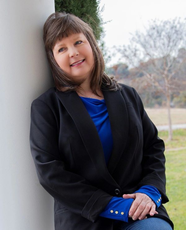 Smiling woman in a black blazer and blue shirt leaning against a column outdoors.