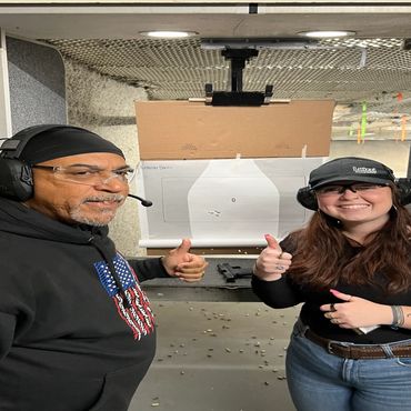 Two people giving thumbs up at an indoor shooting range with target behind.