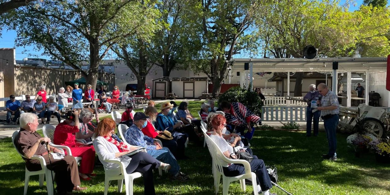 Group of elderly people seated outdoors listening to a speaker under trees.