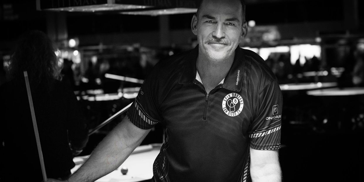 Smiling man in a Spin Doctors shirt leans on a pool table in a dimly lit room.