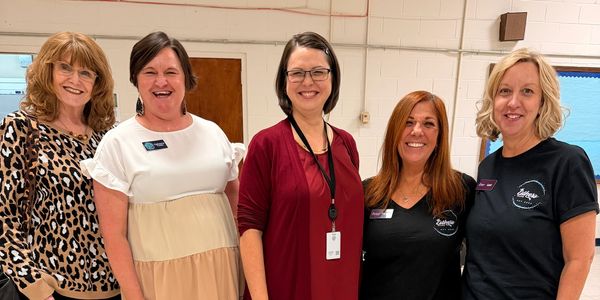 Five women smiling indoors, casually dressed and standing side by side.