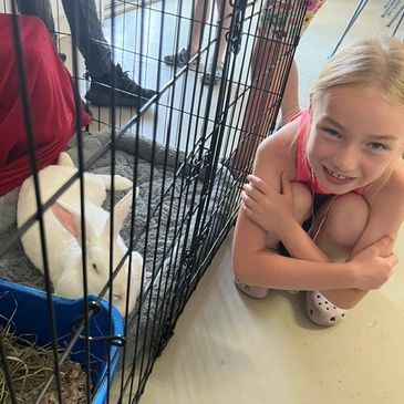 A smiling girl crouches beside a white rabbit in a cage.