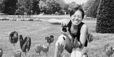 A woman of Asian descent is crouching behind some tulips with a camera in her hands and a big smile
